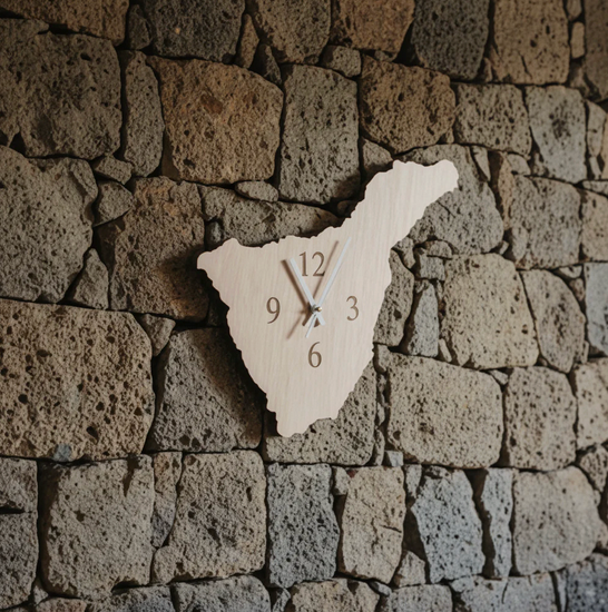 Reloj de pared con forma de Tenerife en madera clara, montado en pared de piedra volcánica – artesanía de Canarias